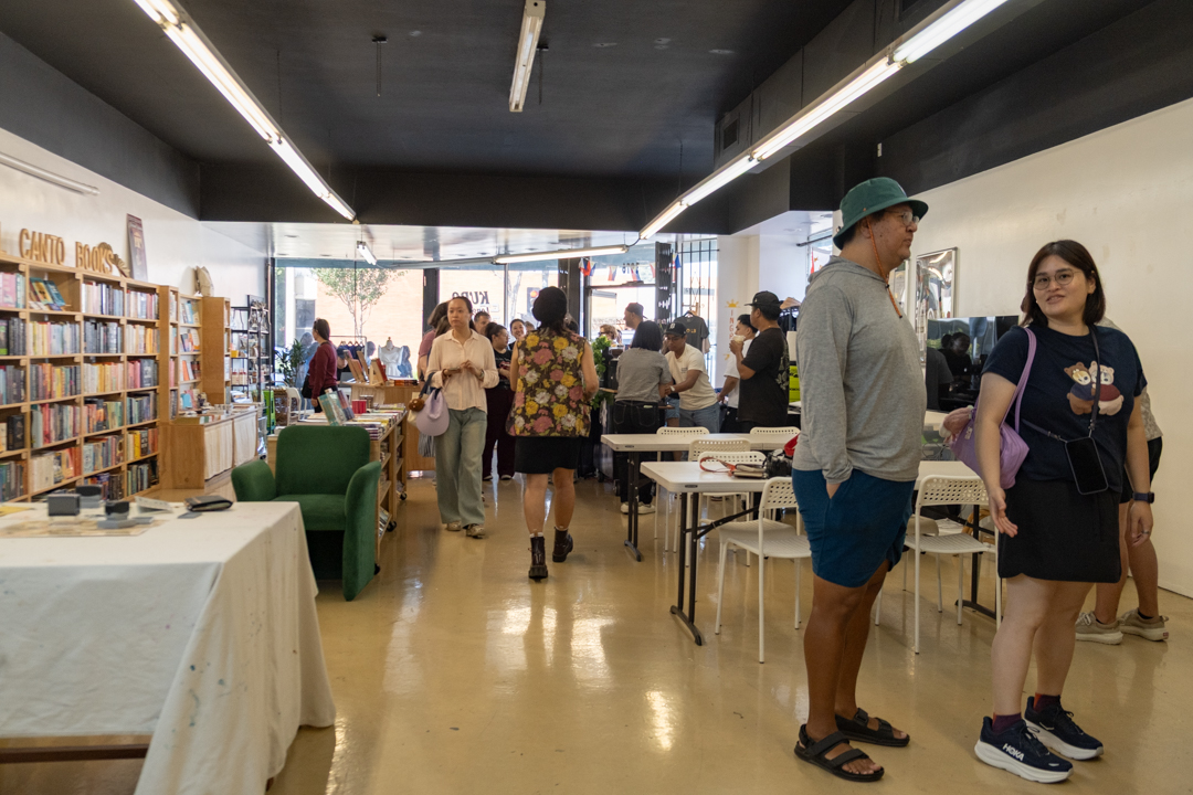 People browse a cozy "Bel Canto Books: bookstore, with shelves filled with books on the left and a lively crowd gathered in the center under bright lights. The atmosphere is vibrant and engaging.