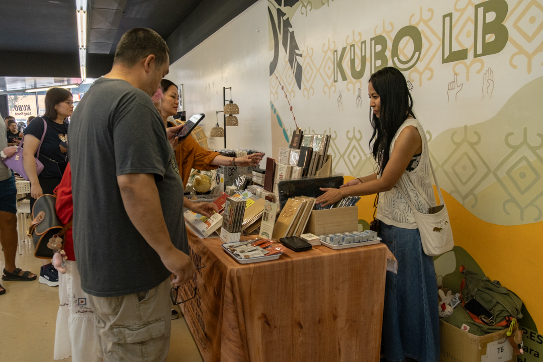 People browse the TRC USA booth at KUBO LB, featuring books, paper goods and stationery on the vendor table. The atmosphere is lively and engaging.