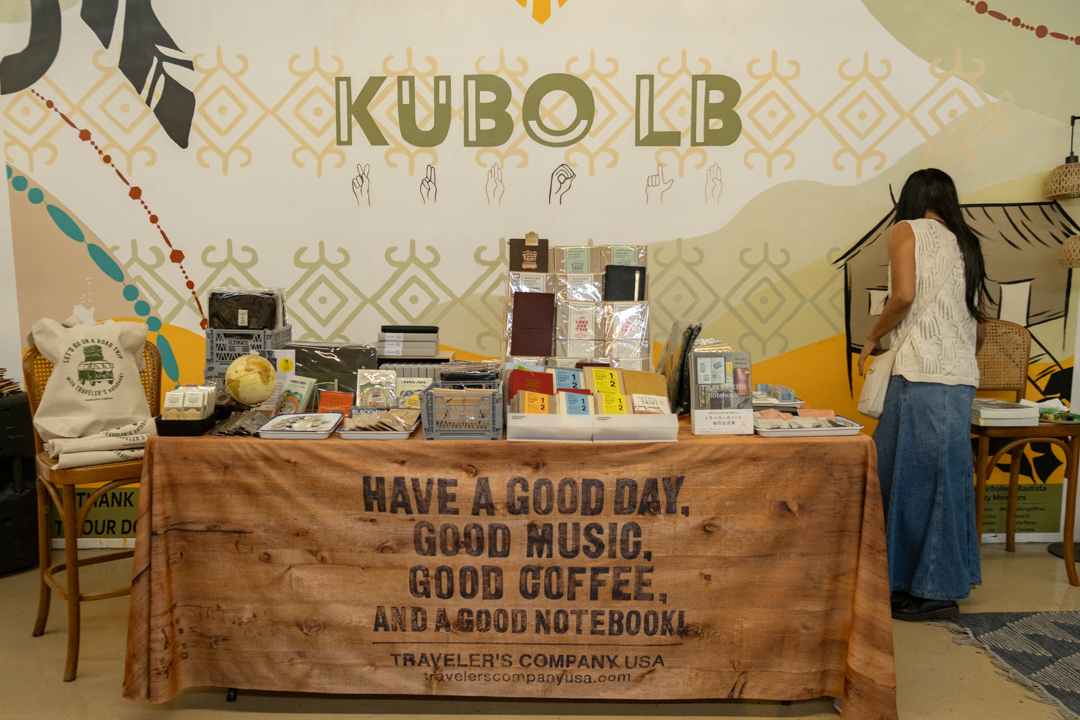 A market stall with a variety of notebooks and stationery displayed on a table covered with a brown cloth that reads, "Have a good day, good music, good coffee, and a good notebook." A person with long hair stands to the right. The background features a decorative wall with "KUBO LB" and geometric patterns. The atmosphere is cozy and inviting.