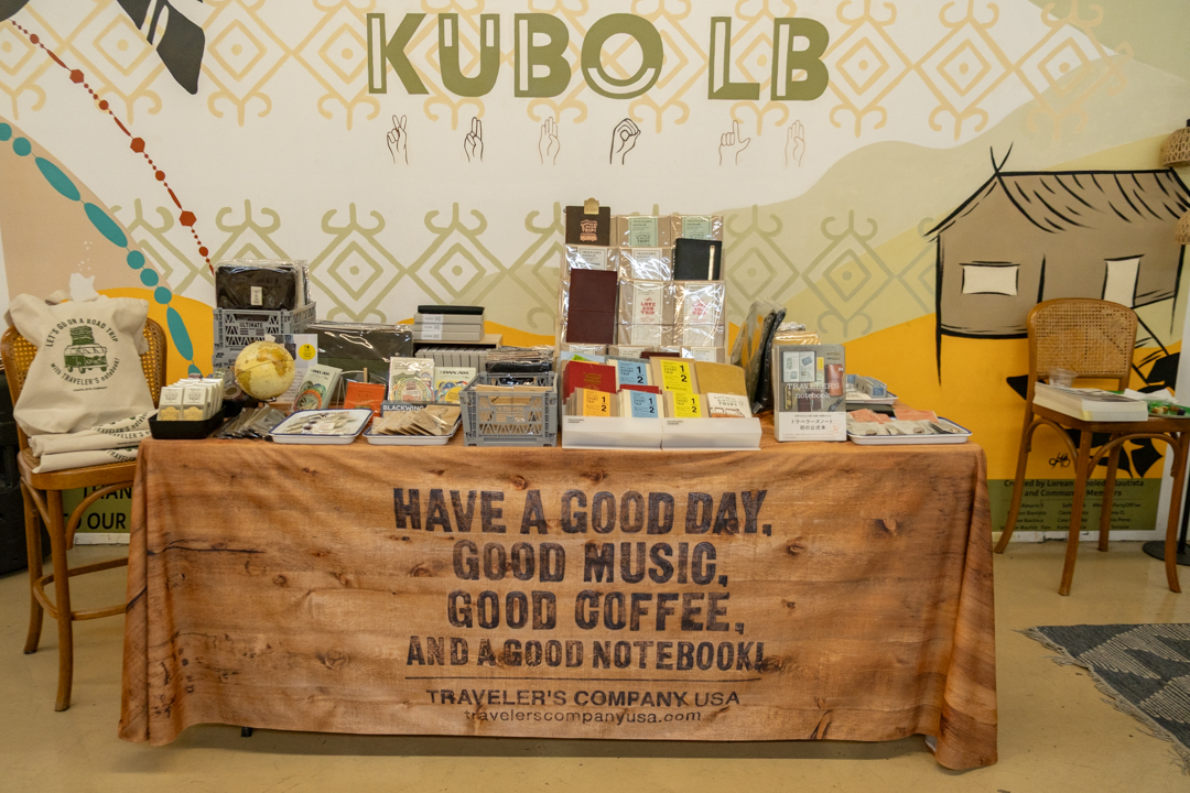 A TRC USA market stall with a variety of notebooks and stationery displayed on a table covered with a brown cloth that reads, "Have a good day, good music, good coffee, and a good notebook." A person with long hair stands to the right. The background features a decorative wall with "KUBO LB" and geometric patterns. The atmosphere is cozy and inviting.