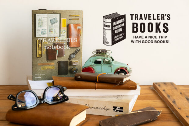 The TRAVELER'S GUIDE book propped up against other books and TRAVELER'S notebooks on a wooden table.