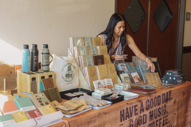 A person restocking products on the TRC USA table filled with TRAVELER'S COMPANY and TRAVELER'S FACTORY Original items, at the SF Pen Show.