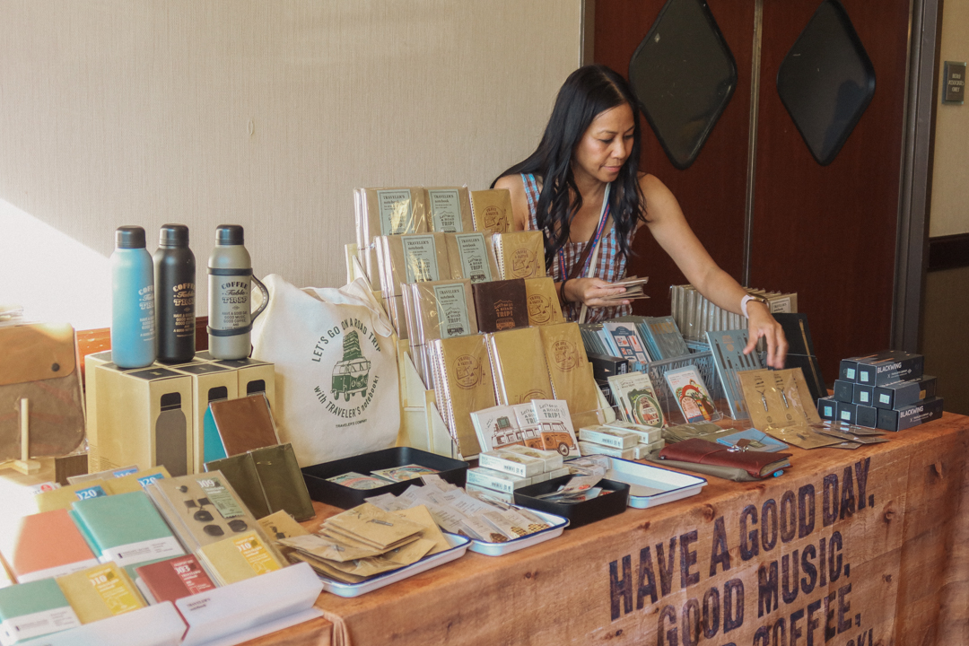A person restocking products on the TRC USA table filled with TRAVELER'S COMPANY and TRAVELER'S FACTORY Original items, at the SF Pen Show.