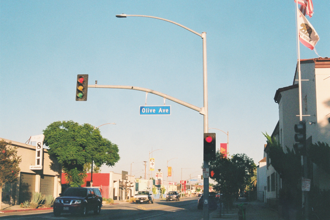 Street view of Olive Ave intersection under a clear blue sky. Traffic lights are red, cars wait below. A tree and buildings line the street. Urban and calm setting.