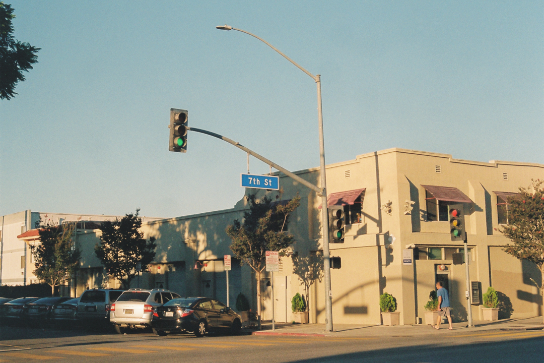 A corner building at 7th Street in warm sunlight. A man walks past, and cars are parked along the street. Trees cast shadows, creating a serene atmosphere.