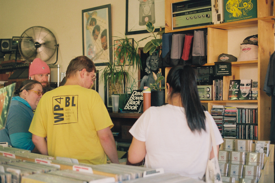 People browse records in a cozy, vintage-style music store with wood shelves, posters, and various albums. The mood is relaxed and nostalgic.