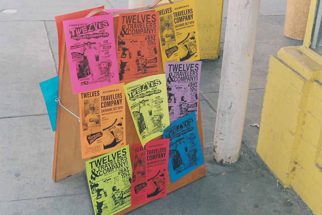 A wooden signboard on a sidewalk displays colorful flyers for "Twelves x Travelers Company Pop-Up" on pink, orange, yellow, green, and blue paper. Bright, vibrant setting.