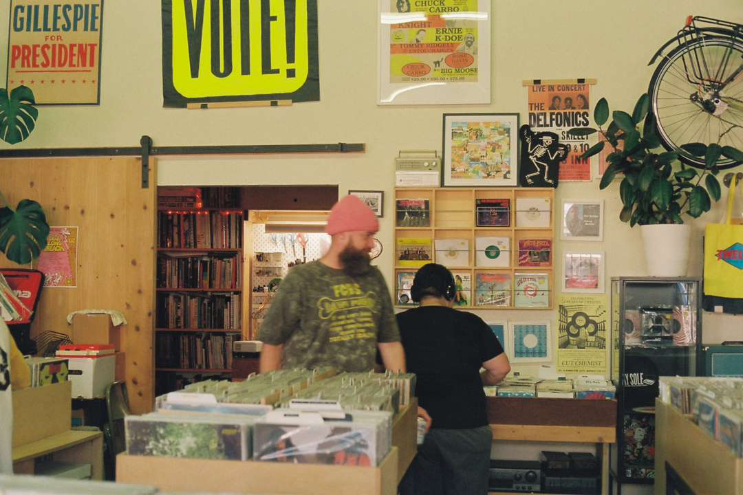 A cozy record store with two people browsing vinyl. Walls are adorned with colorful posters, plants, and a bicycle. A warm, nostalgic atmosphere prevails.