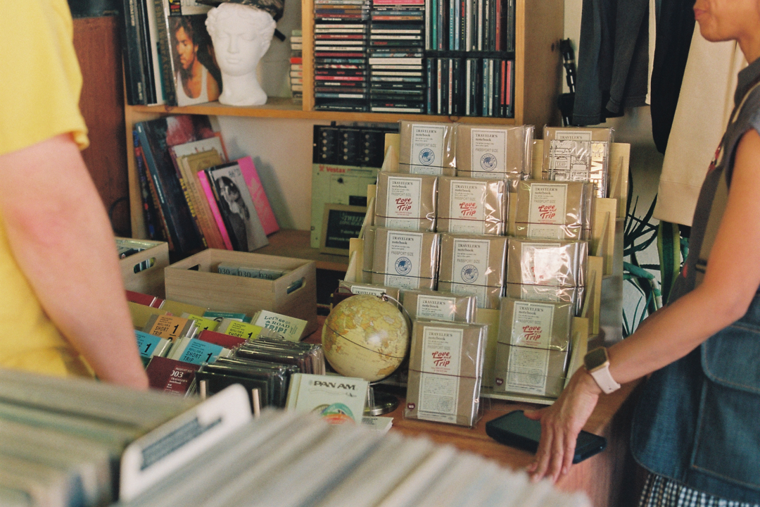 Two people browse colorful records and notebooks on a wooden table in a cozy store. A small globe and stacked items add a vintage feel.