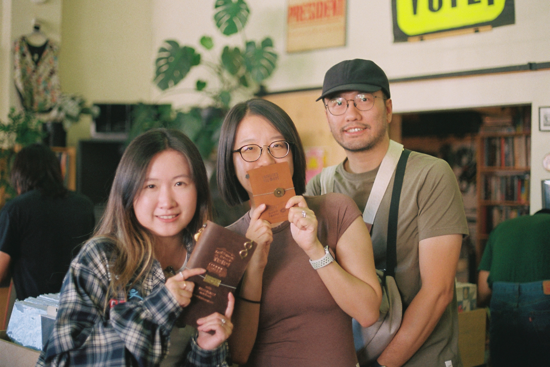 Three people smiling inside a cozy store. Two women hold up small brown notebooks, and a man in glasses and a cap stands beside them. The atmosphere is warm and friendly, with plants and shelves in the background.