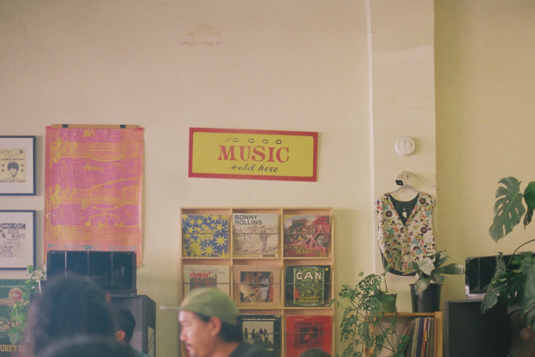 A cozy music shop interior with a "Good Music Sold Here" sign. Vinyl records are neatly displayed on shelves. Lush plants and a mannequin add a relaxed vibe.