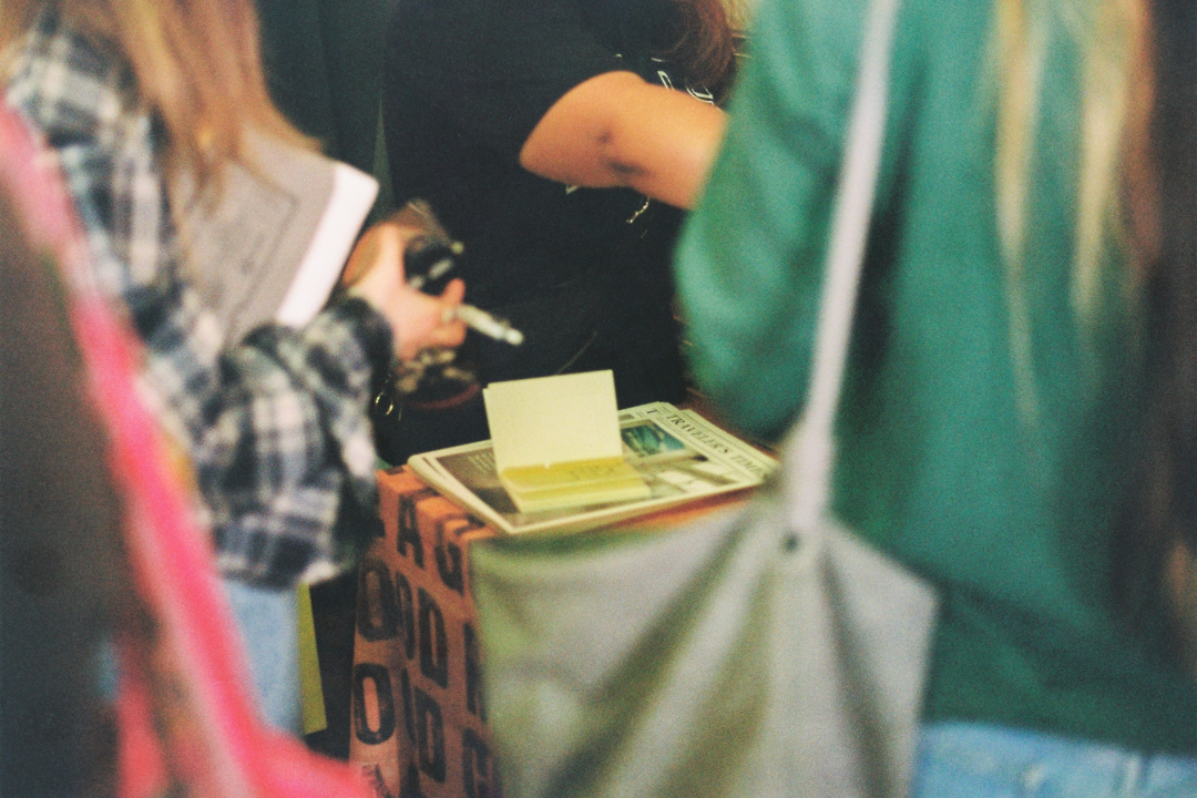 A group of people gather around a table covered with papers and a box. One person holds a notebook, suggesting a casual, social setting.