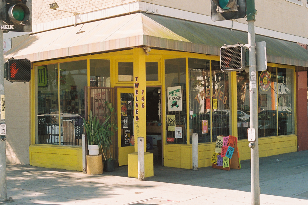 A vibrant, yellow corner storefront of "Twelves Records" with large windows displaying posters and plants. The awning is striped, and the scene is bathed in sunlight, conveying a lively, welcoming atmosphere.
