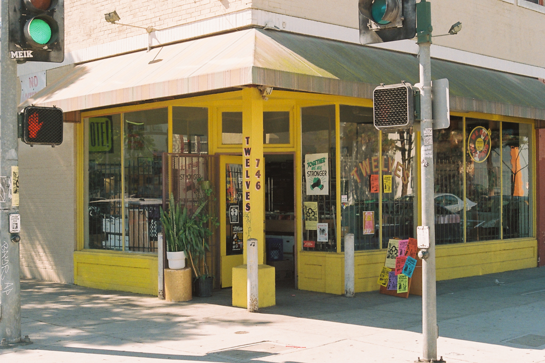 A vibrant, yellow corner storefront with large windows displaying posters and plants. The awning is striped, and the scene is bathed in sunlight, conveying a lively, welcoming atmosphere.