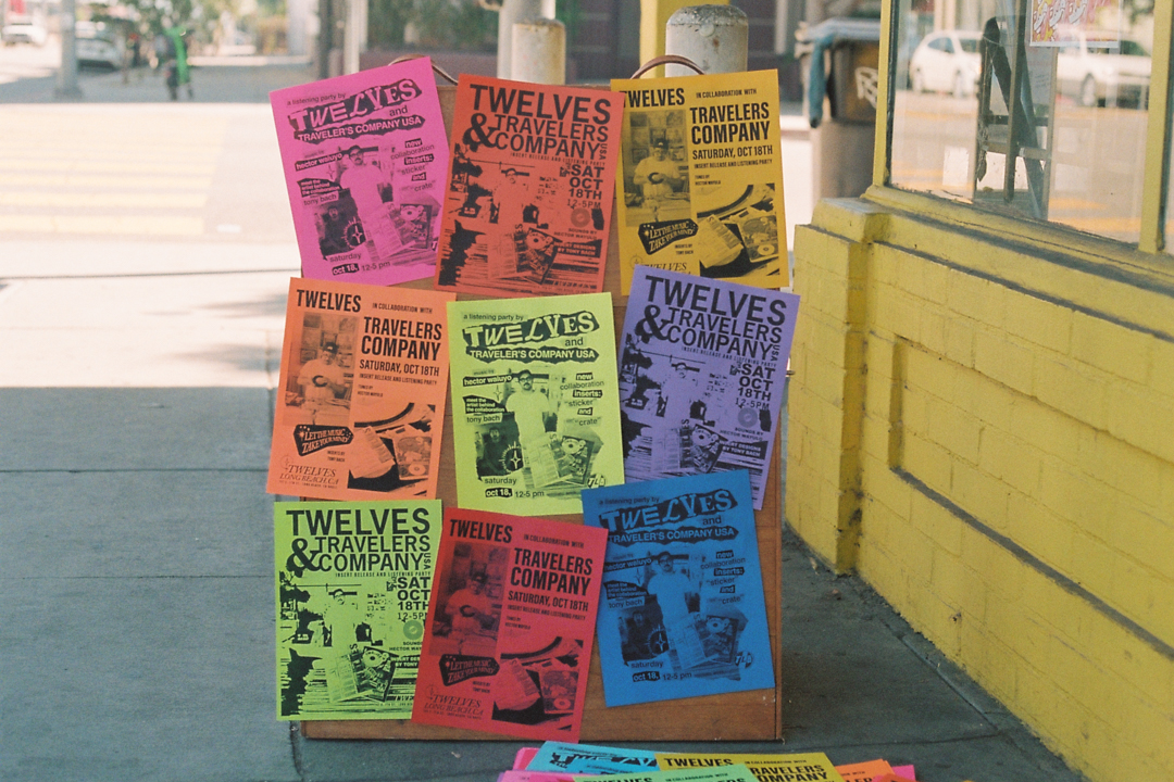 A wooden signboard on a sidewalk displays colorful flyers for "Twelves x Travelers Company Pop-Up" on pink, orange, yellow, green, and blue paper. Bright, vibrant setting.