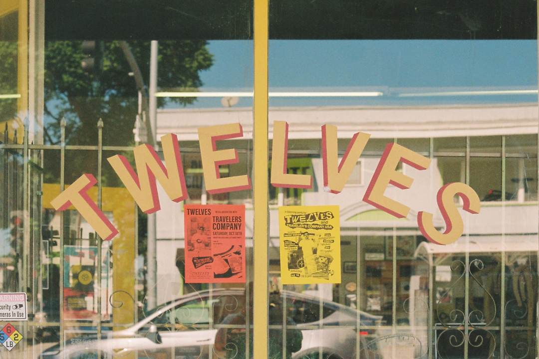 Shop window with the word "TWELVES" in large letters. Two flyers, one red and one yellow, are displayed. A car and buildings reflect outside.