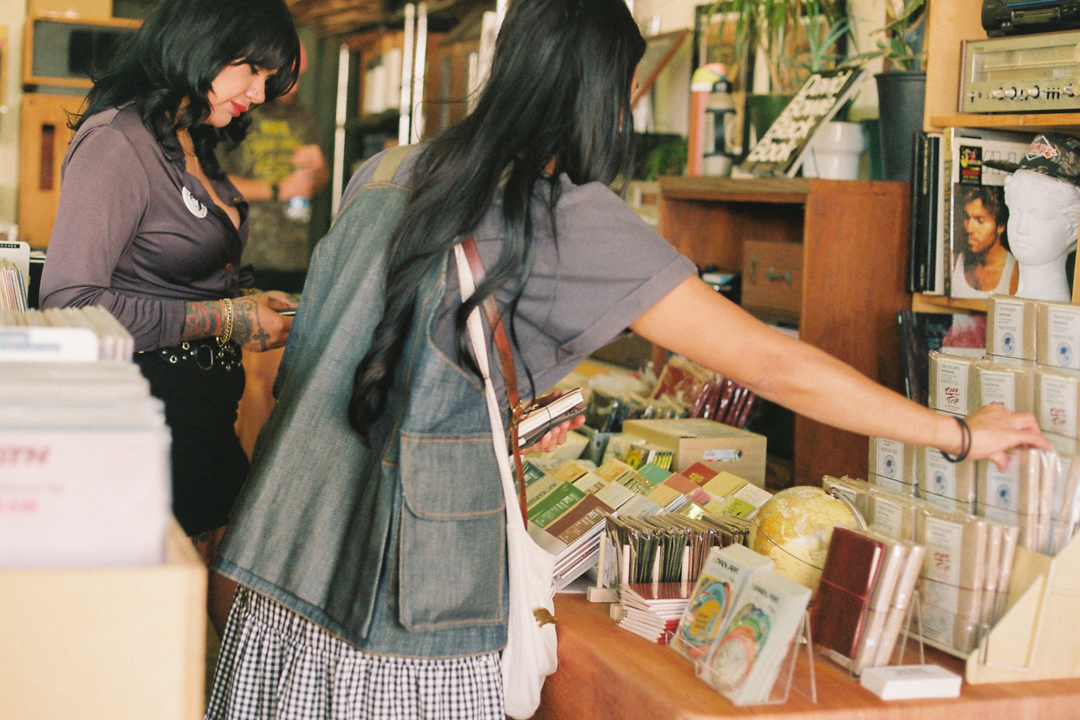 Two women browse in a cozy shop filled with books, CDs, TRAVELER'S notebooks, and stationery. The atmosphere is warm and inviting, with potted plants on shelves.