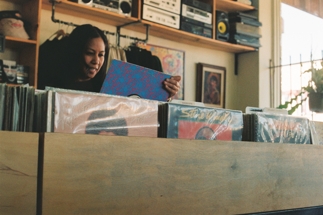 A person in a record store flipping through vinyls. They're holding a blue record cover with pink text. Shelves with decor and music equipment are in the background, creating a nostalgic, relaxed atmosphere.