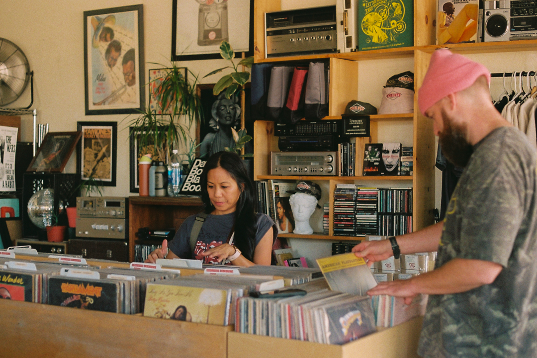 Two people browse vinyl records in a music store filled with albums, posters, plants, and audio equipment.
