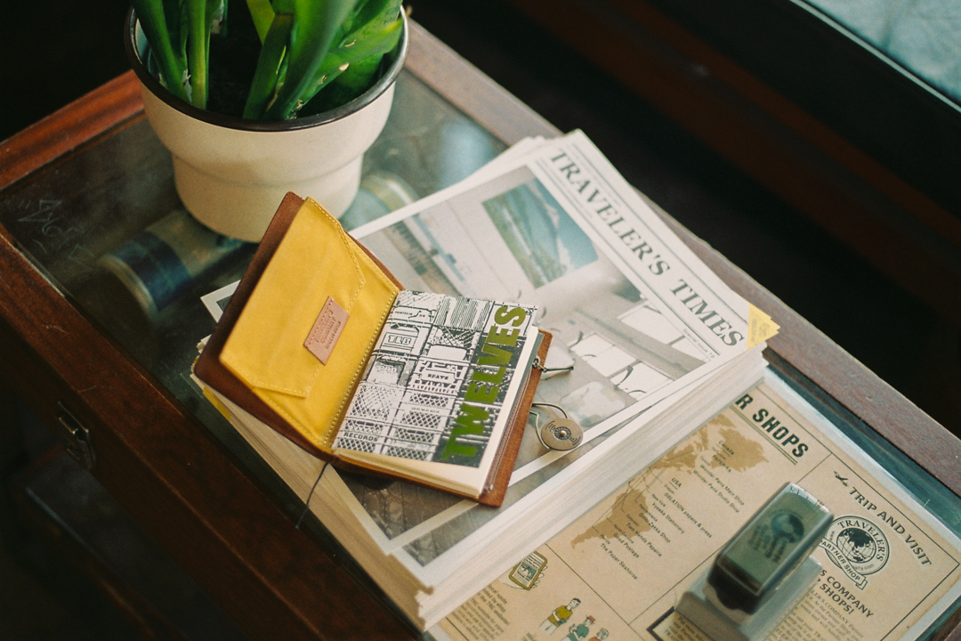 A potted plant sits on a glass table beside a stack of Traveler's Times newspapers. A map, a stamp, and an open leather wallet with a colorful design are visible. The scene conveys a vintage, travel-inspired vibe.