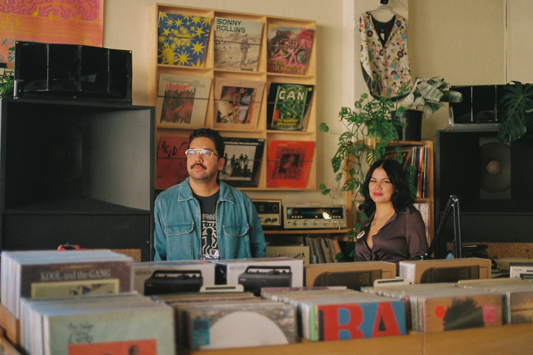 Two people stand in a vibrant record store, surrounded by vinyl records and plants. The atmosphere is nostalgic and inviting, with colorful album covers on the wall behind them.