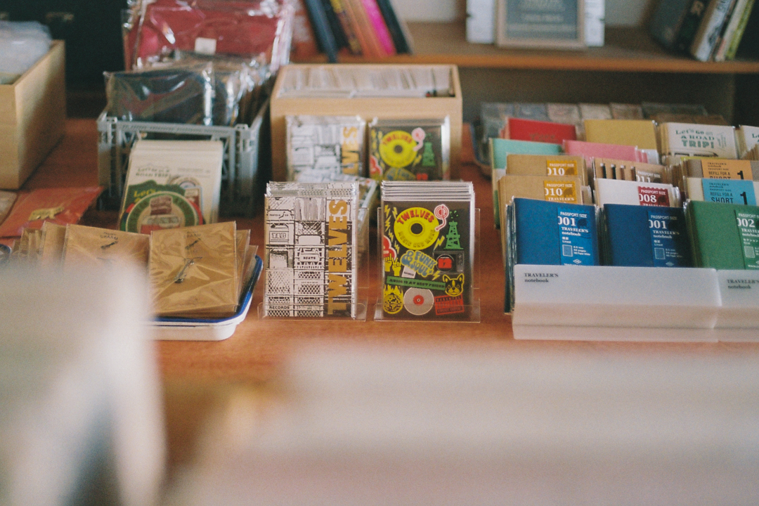 A tidy store display features various stationery items, including colorful "Crate" and "Sticker" collaboration notebooks and postcards, set on a wooden table with a cozy, inviting atmosphere.