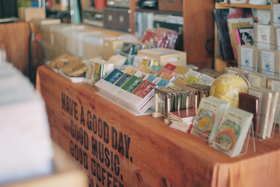 A cozy shop corner featuring a wooden counter with the text "Have a good day. Good music, good coffee." Displayed are colorful booklets, CDs, and a small globe, creating a warm, inviting atmosphere.