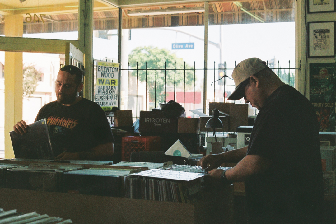 Two men browse vinyl records in a cozy record shop with sunlit windows. One wears a black T-shirt, the other a cap, both focused and relaxed.