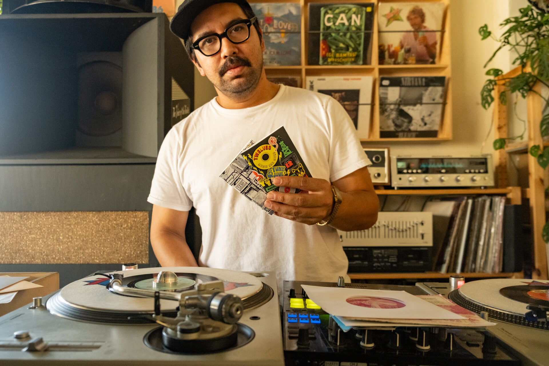 Shop owner Hector behind a DJ booth at TWELVES RECORD, holding the TWELVES X TRC notebook inserts.