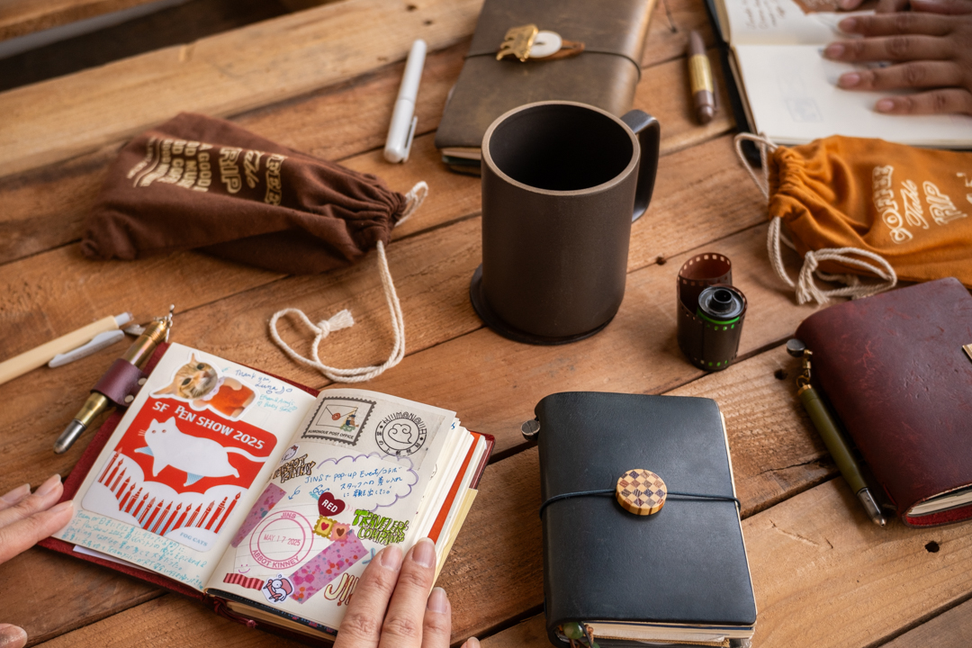 A collection of TRAVELER'S notebooks and COFFEE TABLE TRIP tools and two people's hands around the table, enjoying conversation over their notebooks.