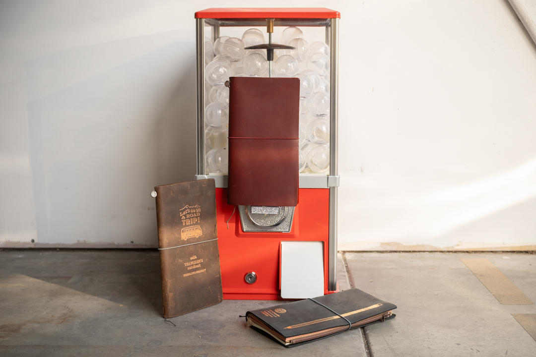 Special Gachapon event featuring a red gachapon machine, with three TRAVELER'S notebooks displayed next to it.