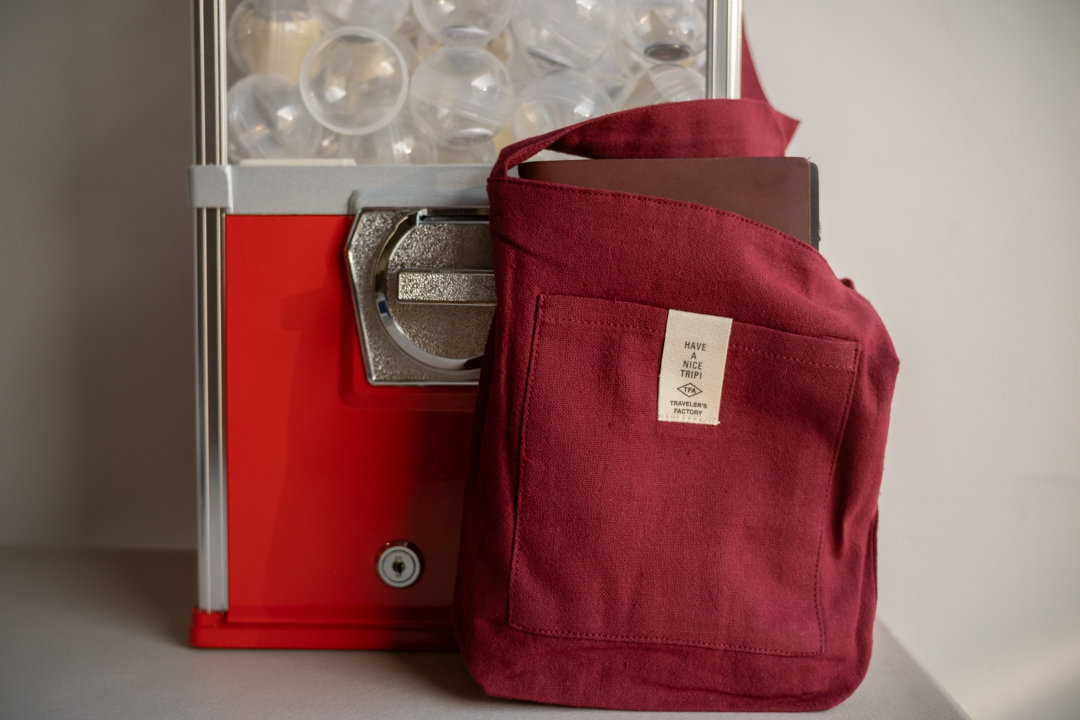 A red sacoche with a TRAVELER'S notebook in brown inside the bag, resting against a red gachapon machine.