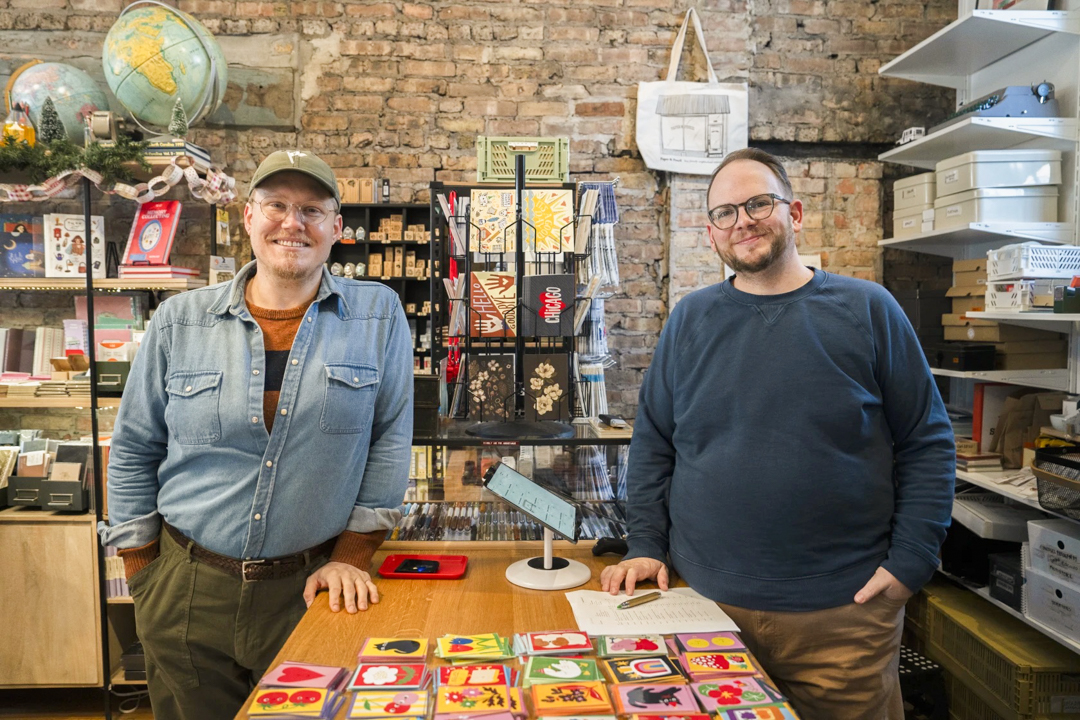Tyler and Eric, the owners of Paper & Pencil, are standing near the register of their own stationery shop.