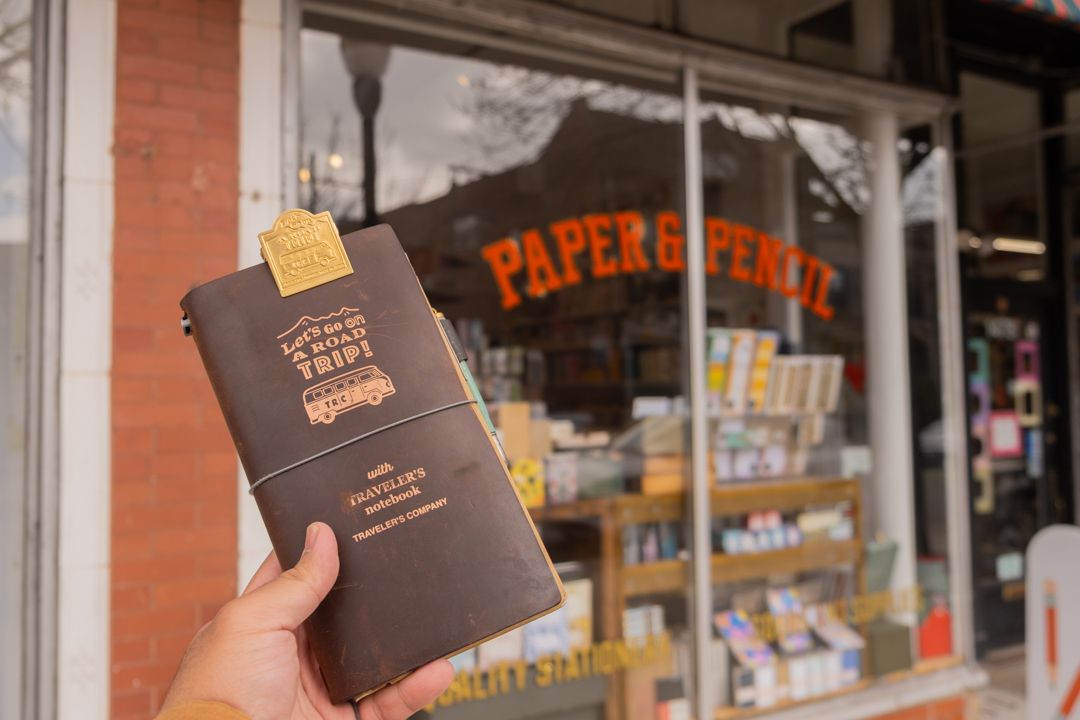 A person holding a TRAVELER'S notebook outside of Paper & Pencil store, which features a brick facade, big windows, a sign-painted store name, and a multi-colored awning.