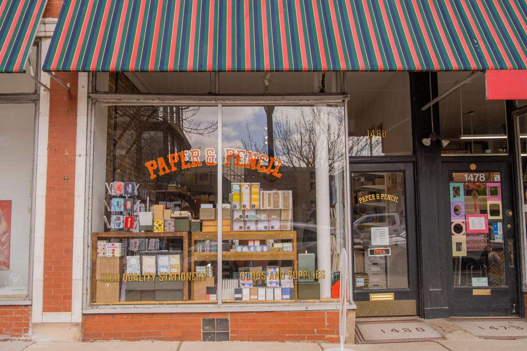 The exterior of Paper & Pencil, a stationery store located in Andersonville, Chicago, which has a brick facade, big windows, a sign-painted store name, and a multi-colored awning.