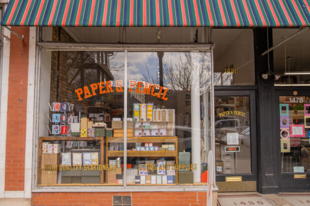 The exterior of Paper & Pencil, a stationery store located in Andersonville, Chicago, which has a brick facade, big windows, a sign-painted store name, and a multi-colored awning.