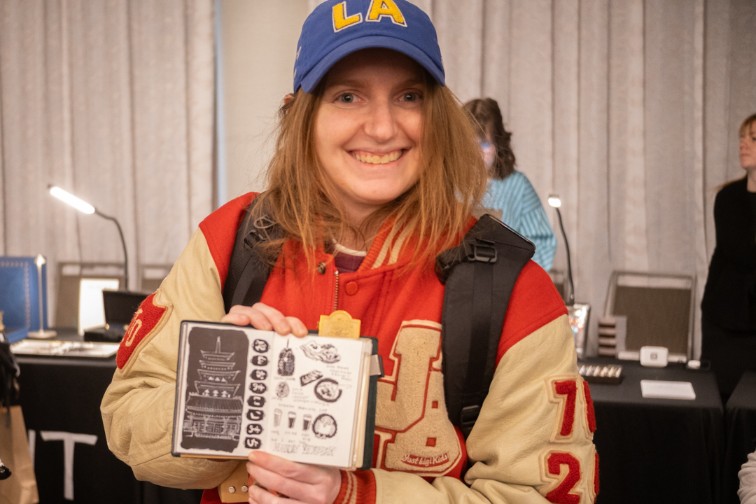 A smiling woman showcasing her drawings in her passport size TRAVELER'S notebook.