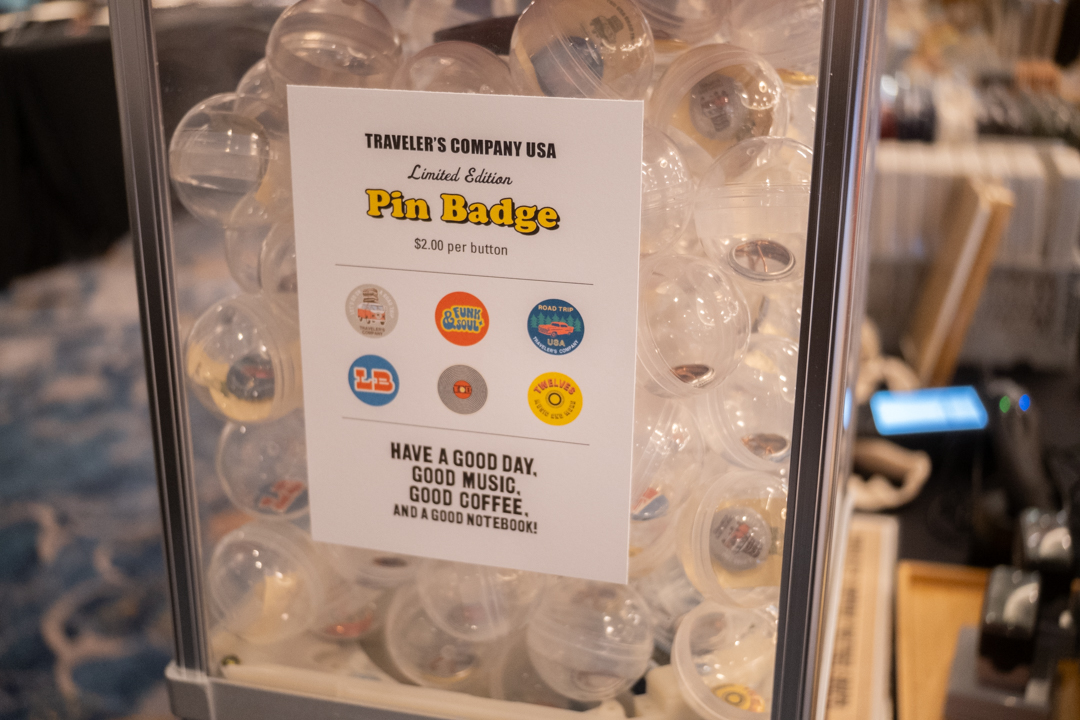 A close-up of the capsule toy machine filled with pin buttons resting on the TRAVELER'S COMPANY USA table at the California pen show.