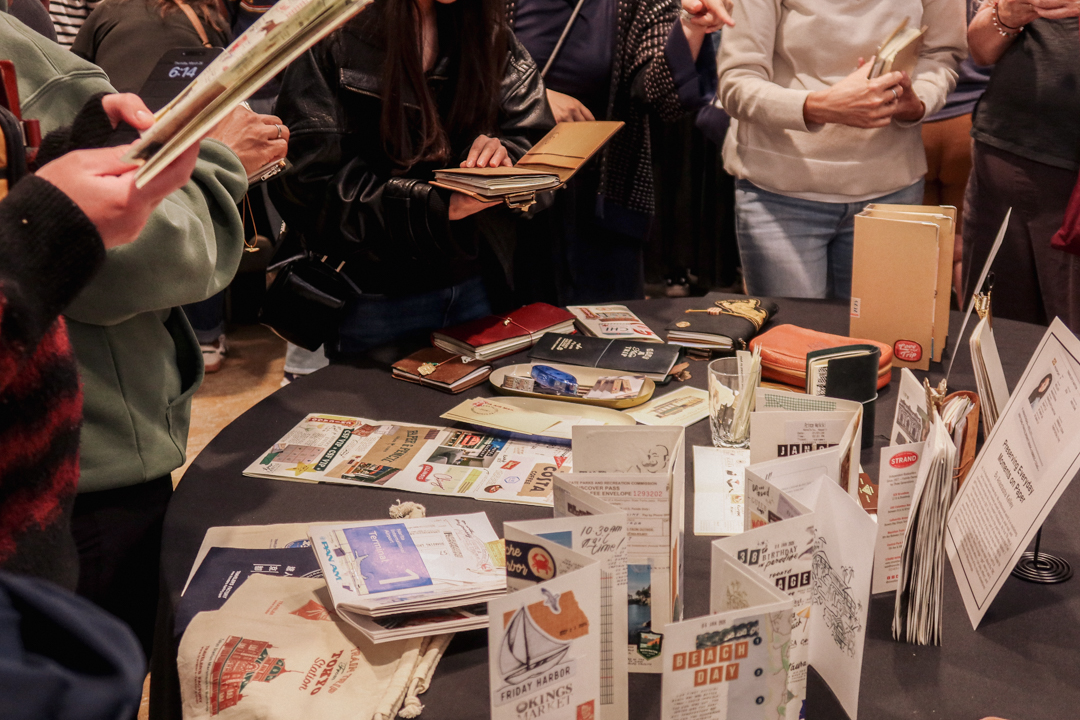People are surrounding a round table, which is an exhibit of travel journals created by artists who use TRAVELER'S notebooks.