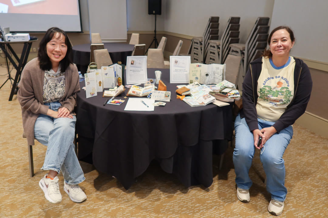Two artists sitting beside their exhibit of travel journals and TRAVELER'S notebooks items, which are on a round table with a black tablecloth.