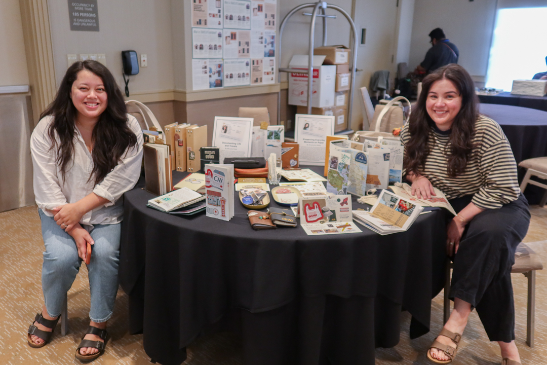 Two artists sitting beside their exhibit of travel journals and TRAVELER'S notebooks items, which are on a round table with a black tablecloth.