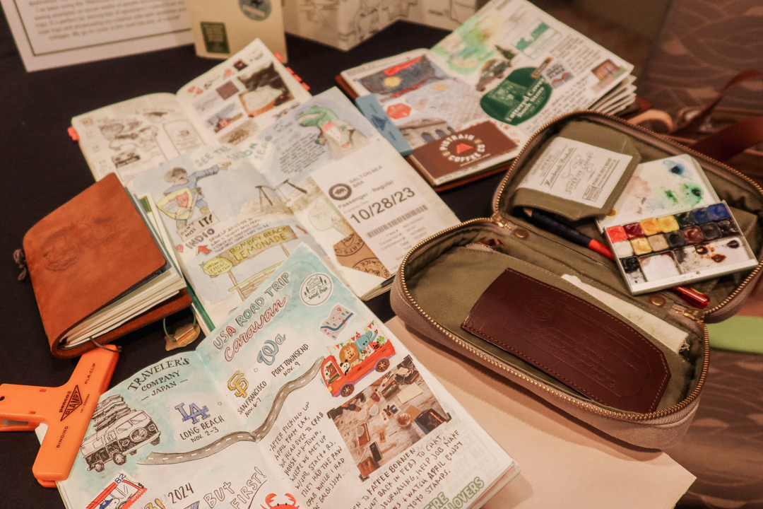 An exhibit of travel journals created by artists who use TRAVELER'S notebooks on a round table with black tablecloth.