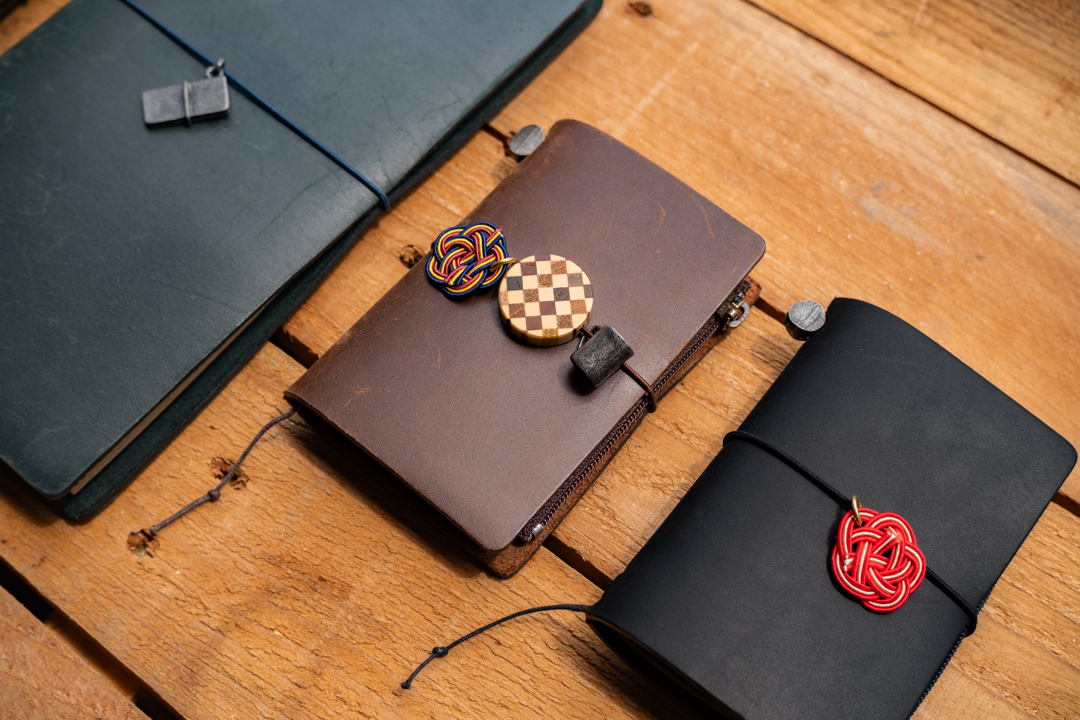 TRAVELER'S notebooks decorated with Tin, YOSEGI, and MIZUHIKI charms against a wooden table.