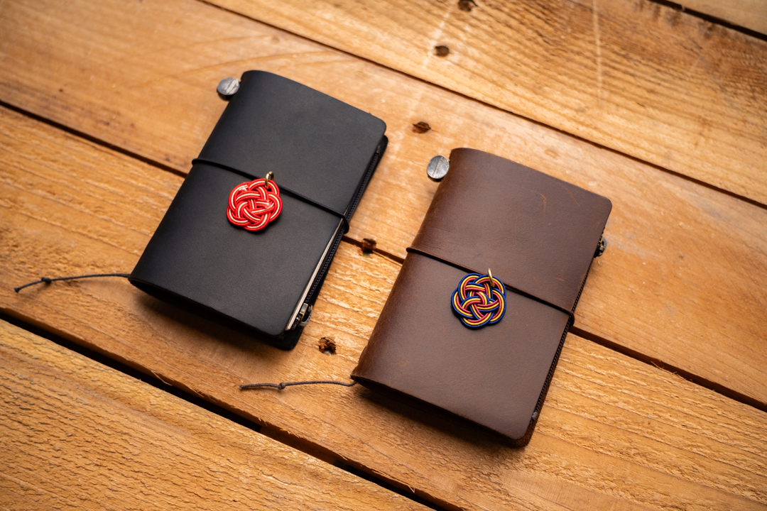 A black and brown TRAVELER'S notebooks in Card Size decorated with MIZUHIKI knot charms, on a wooden table.