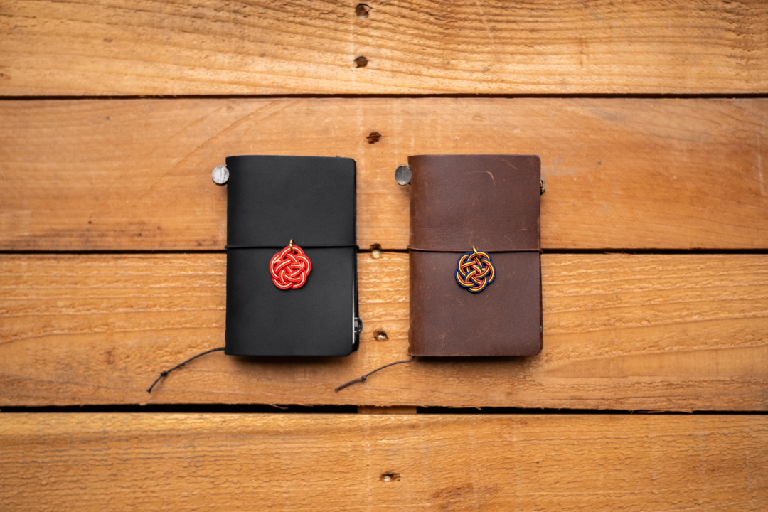 A black and brown TRAVELER'S notebooks in Card Size decorated with MIZUHIKI knot charms, on a wooden table. 