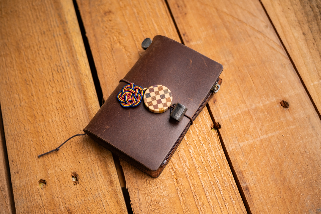 A brown TRAVELER'S notebook Card Size decorated with mug tin charm, YOSEGI wooden charm, and MIZUHIKI knot charm, on a wooden table. 