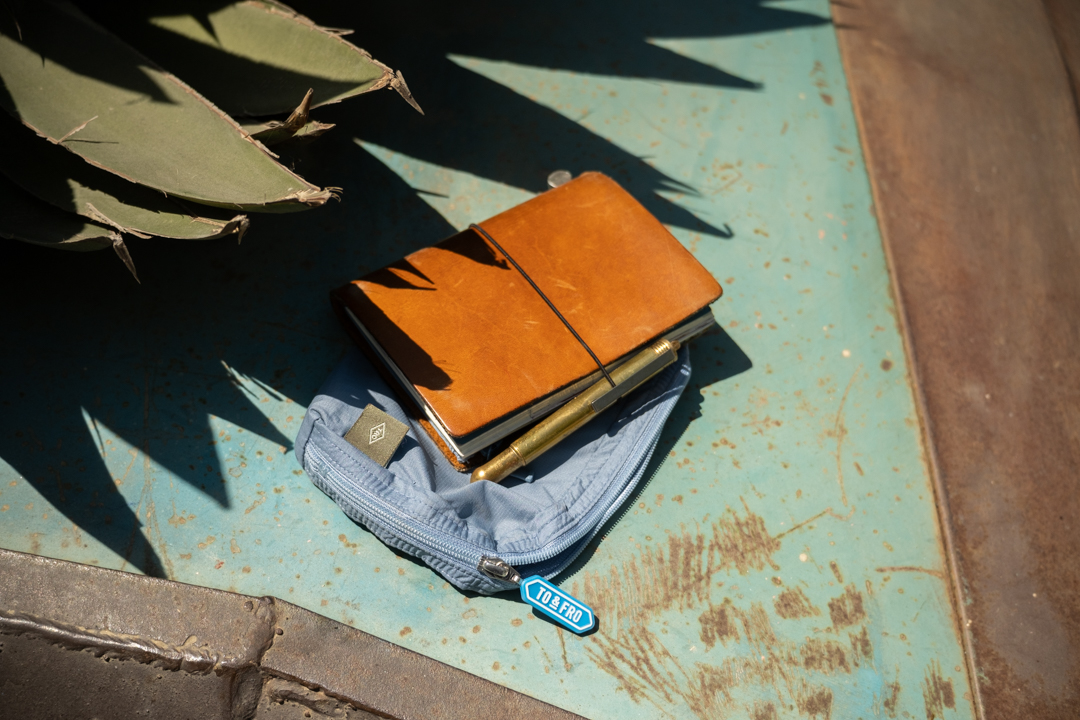 A camel TRAVELER'S notebook in passport size with a blue TO&FRO Organizer beside an agave plant.