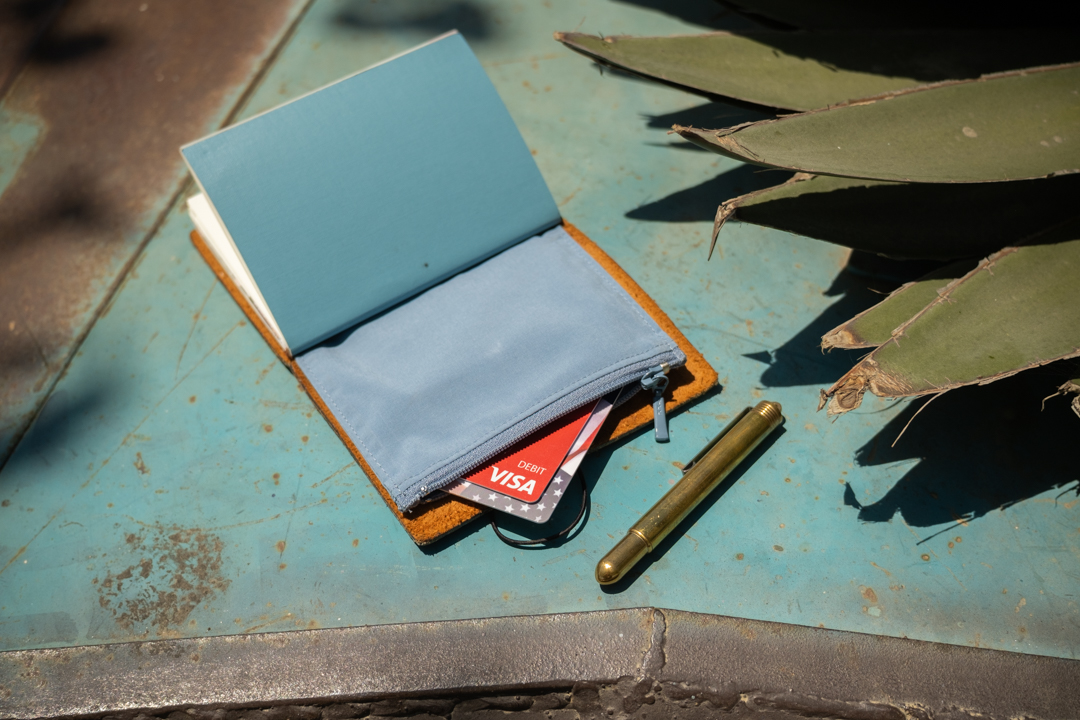 A camel TRAVELER'S notebook open to the TO&FRO zipper case, showing bank cards and a brass pen beside an agave plant.