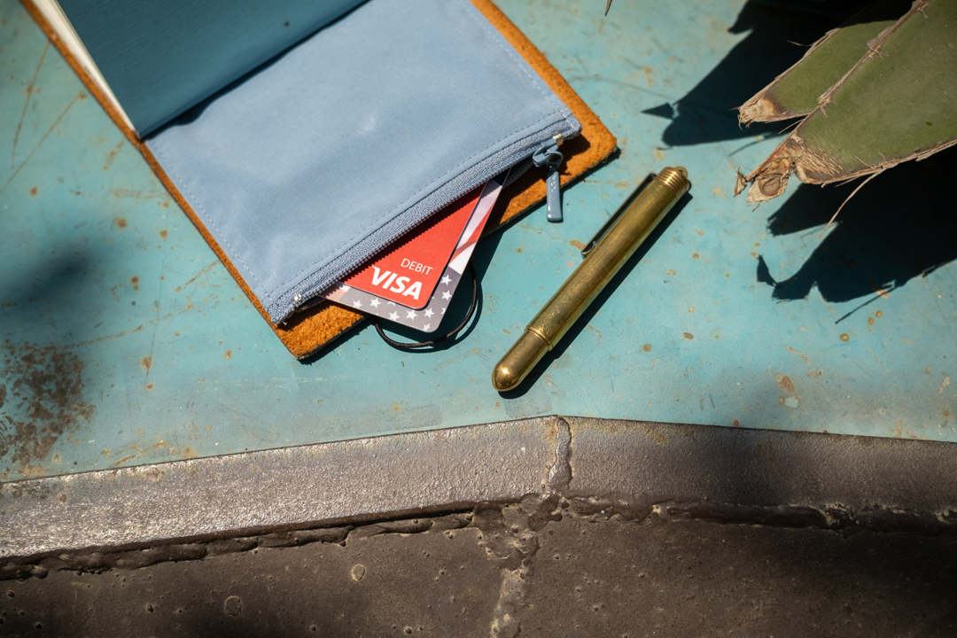 A camel TRAVELER'S notebook open to the TO&FRO zipper case, showing bank cards and a brass pen beside an agave plant.