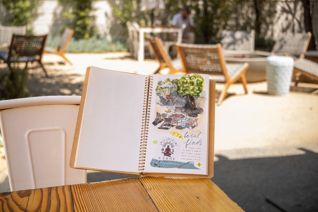 A painting of the outdoor patio in a spiral ring notebook which is propped up a a chair and restaurant table.